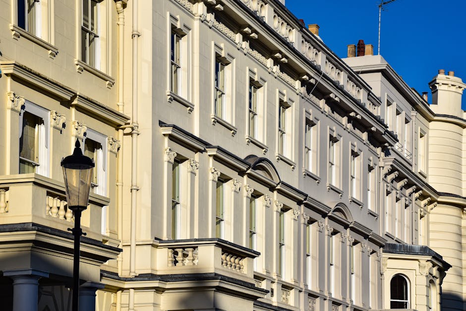 The image depicts a residential street with a row of elegant, white-painted Victorian-style terraced houses featuring decorative columns, black wrought-iron railings, and large windows. The pavement i