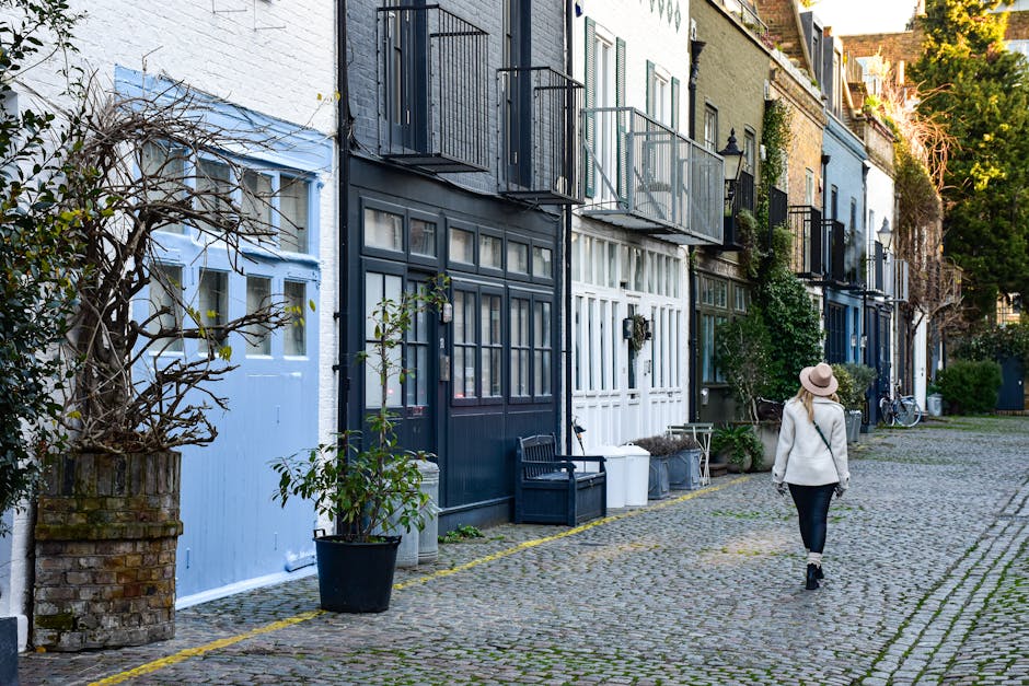 A view of a narrow residential street with tightly packed, traditional Georgian terraced houses featuring brick facades and white window frames, characteristic of Ebury Street mews homes. The image sh