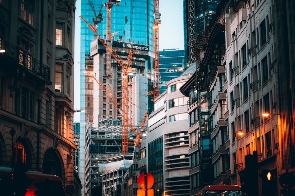 A city street scene depicting a narrow urban road lined with historic and modern buildings, including ornate stone facades and glass-fronted structures. Several pedestrians are walking along the pavem