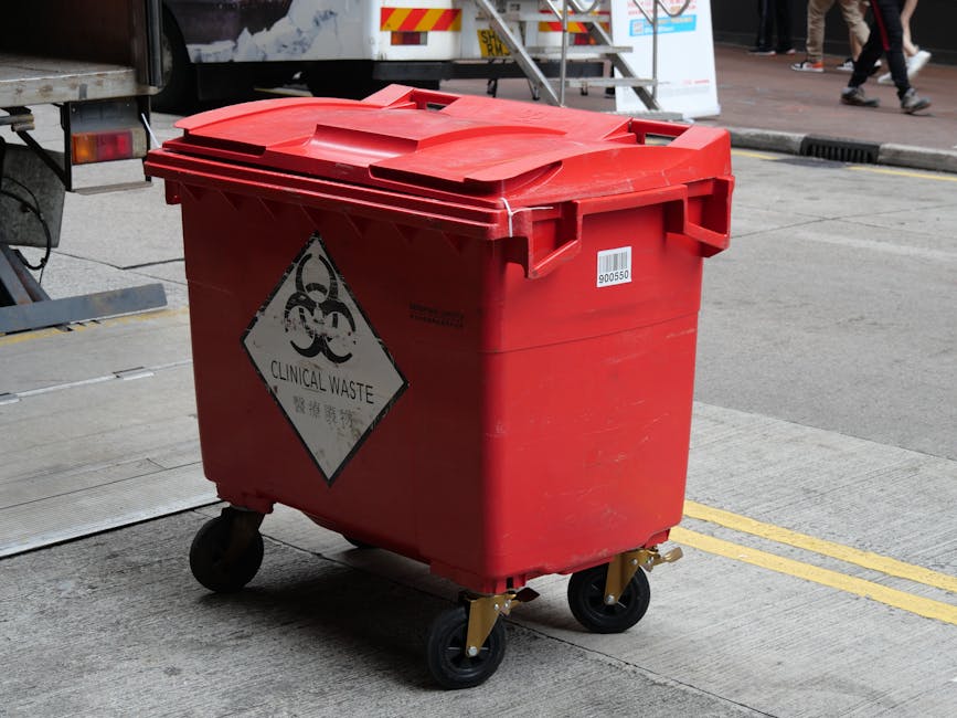 A person’s hand is seen placing a grey disposable coffee cup into a black, open-top recycling container situated outdoors in a garden or green space. The container appears to be made of metal or pla