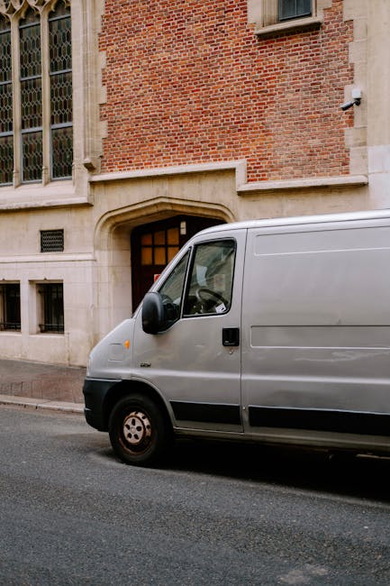 A row of six white commercial vans parked closely together on a paved surface, with visible rear doors and yellow UK license plates. The vans have tinted rear windows, and some feature branding and we