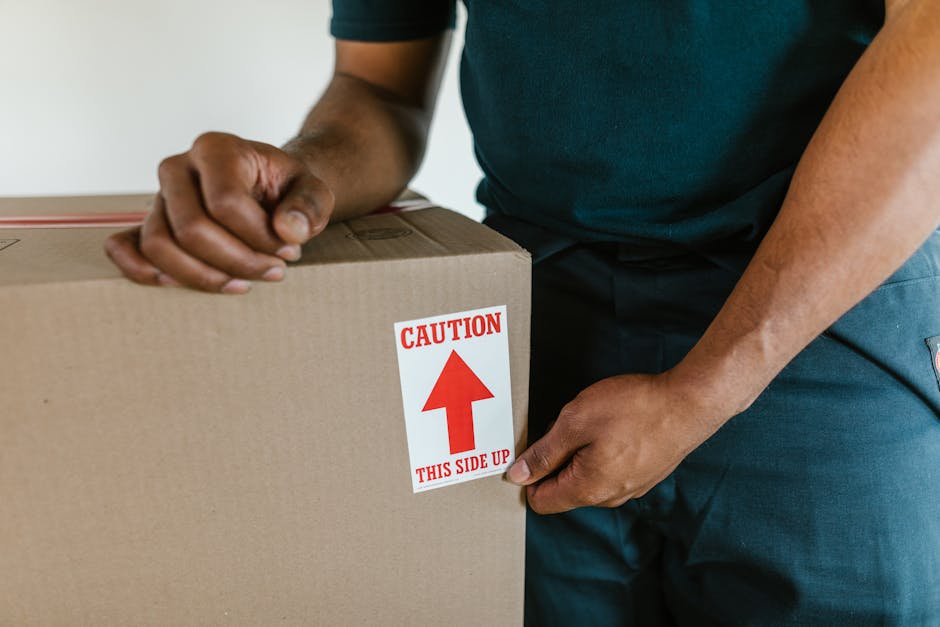 A person wearing a dark green T-shirt and blue work trousers is in the process of moving a large cardboard box, which has a red and white caution label indicating 'Caution: This Side Up' with an upwar